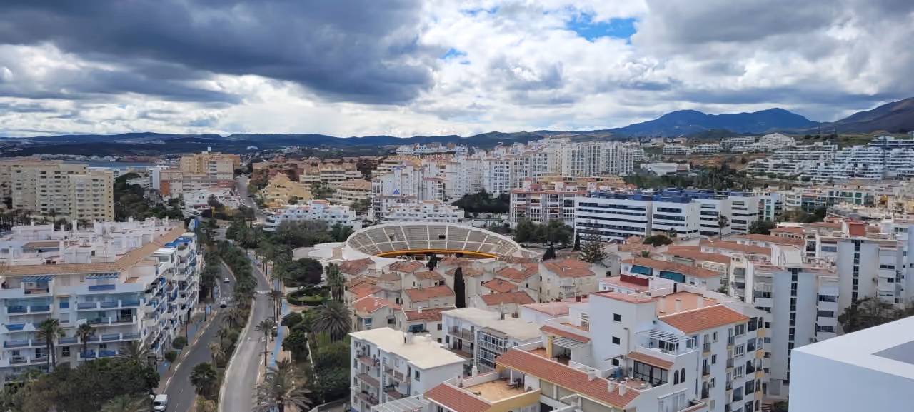 Ophøjet panoramaview over Estepona nær LAS MESAS BLUE HORIZON med tyrefægterarenaen Plaza de Toros, hvidkalkede boligejendomme, en palmekantet boulevard og Sierra Bermeja i baggrunden.