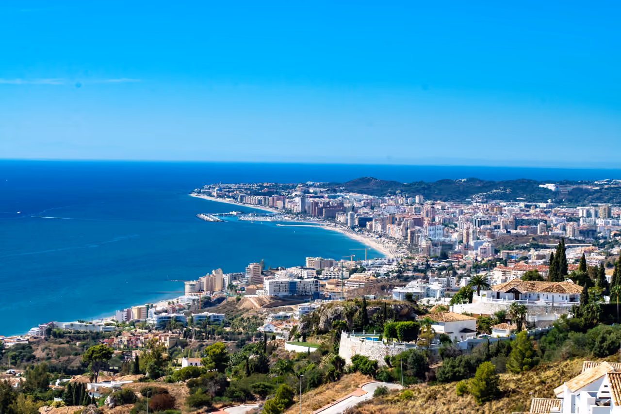 Panoramic elevated coastal view of Fuengirola near CORE HIGUERON FASE II, showing the beach, marina, urban shoreline and Mediterranean Sea, with residential villas on the hillside in the foreground.