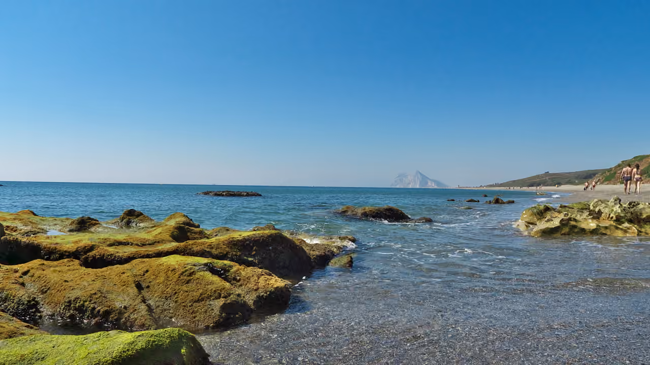 Aluekuva ALTARA ALCAIDESA -kohteen läheltä: Playa de Levanten rannikko lähellä La Línea de la Concepciónia, sammaleisia kallioita, kirkasta Välimeren vettä, hiekkarantaa ja kaukana näkyvä Gibraltarin kallio.