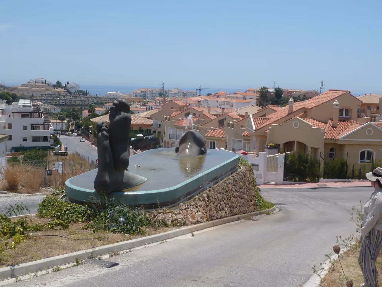 Photo des environs de BOUTIQUE PROJECT montrant une fontaine publique ornée de figures abstraites sur un rond-point, avec le paysage résidentiel à flanc de colline de La Cala de Mijas et la mer Méditerranée en arrière-plan.